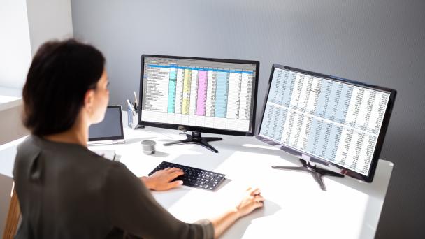 Female looking at two computer screens conducting analysis of spreadsheets