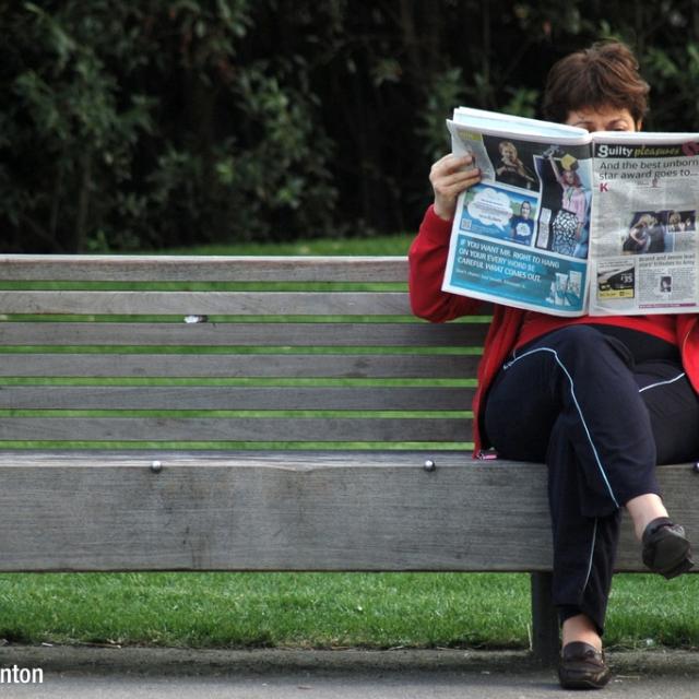 Woman sitting on a bench, reading a newspaper.