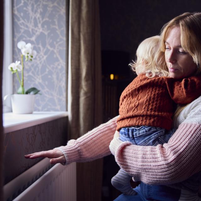 Mother With Son Trying To Keep Warm By Radiator At Home During Cost Of Living Energy Crisis 