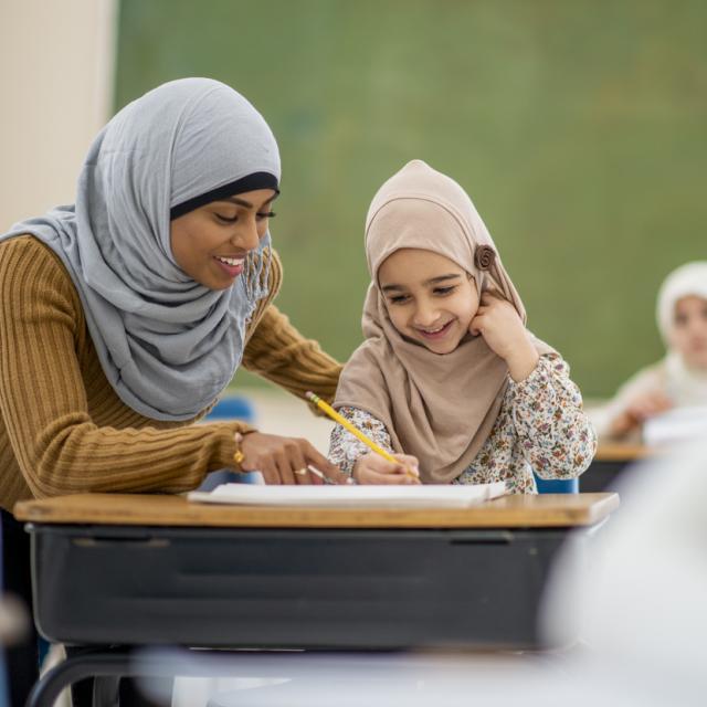 A muslim girl smiles as she sits in class learning. She is looking at her paper that her teacher is pointing at.