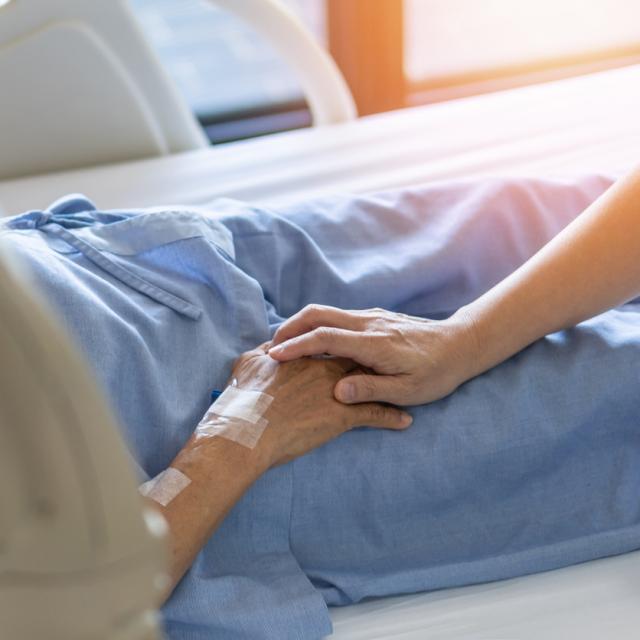 Caregiver holding elderly senior patient (ageing old adult person) hand in hospital bed.