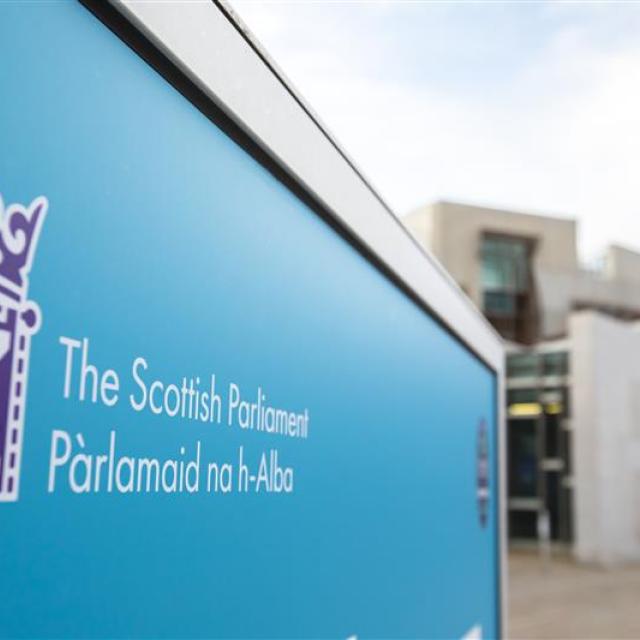 A blue sign to the Scottish Parliament, Parlamaid na h-Alba, is in the foreground and background is blurred buildings with a person walking in front.