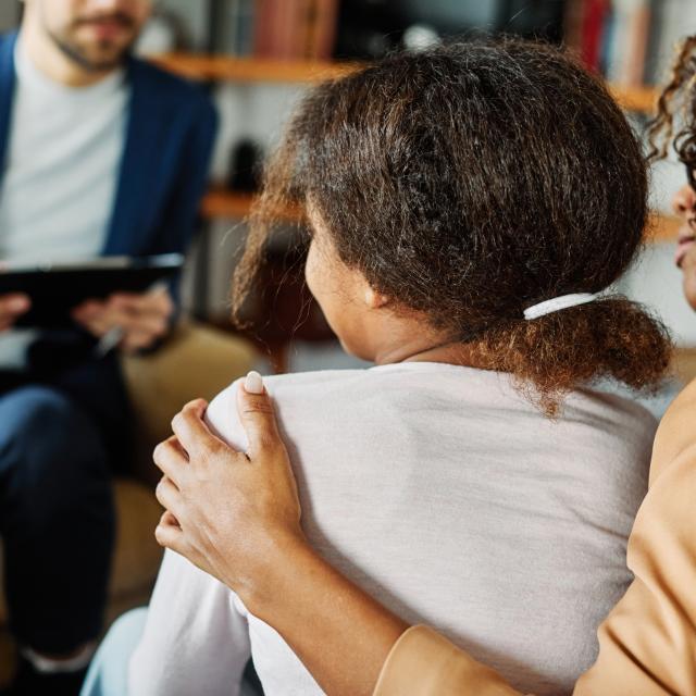 Mother has her arm around her child in the foreground, talking to a therapist, advisor or support worker in the background.