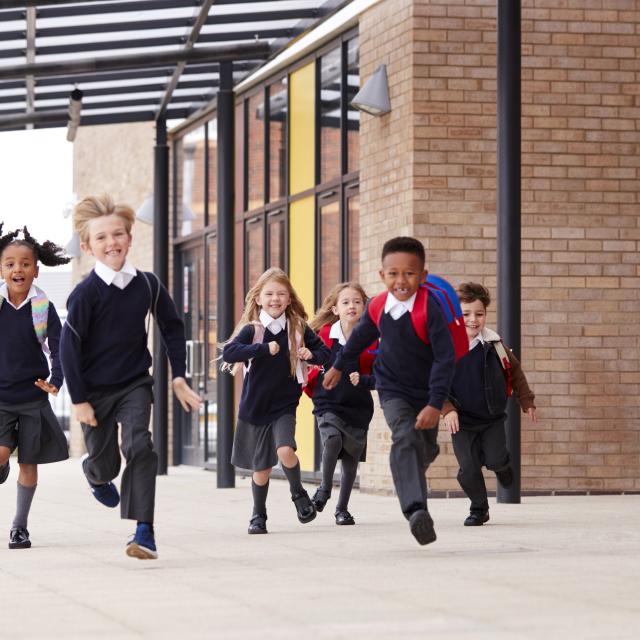 Primary school kids, wearing school uniforms and backpacks, running on a walkway outside their school building, front view