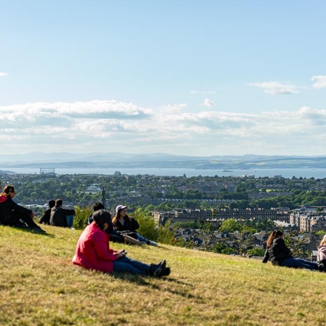 People stretched out on the grass of a mountain hill watching the views of the city of Edinburgh, UK.