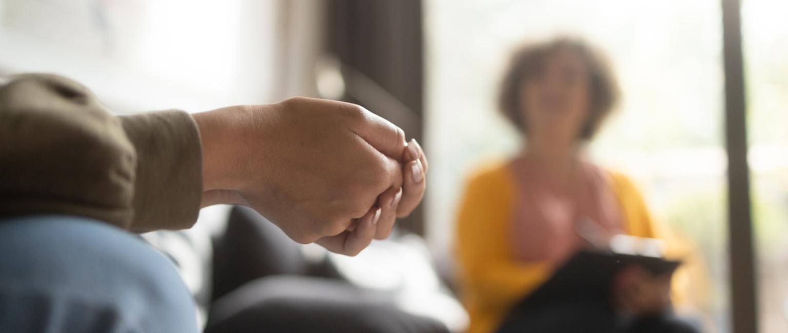Crossed hands in foreground and in focus, in the background a therpaist or interviewer.