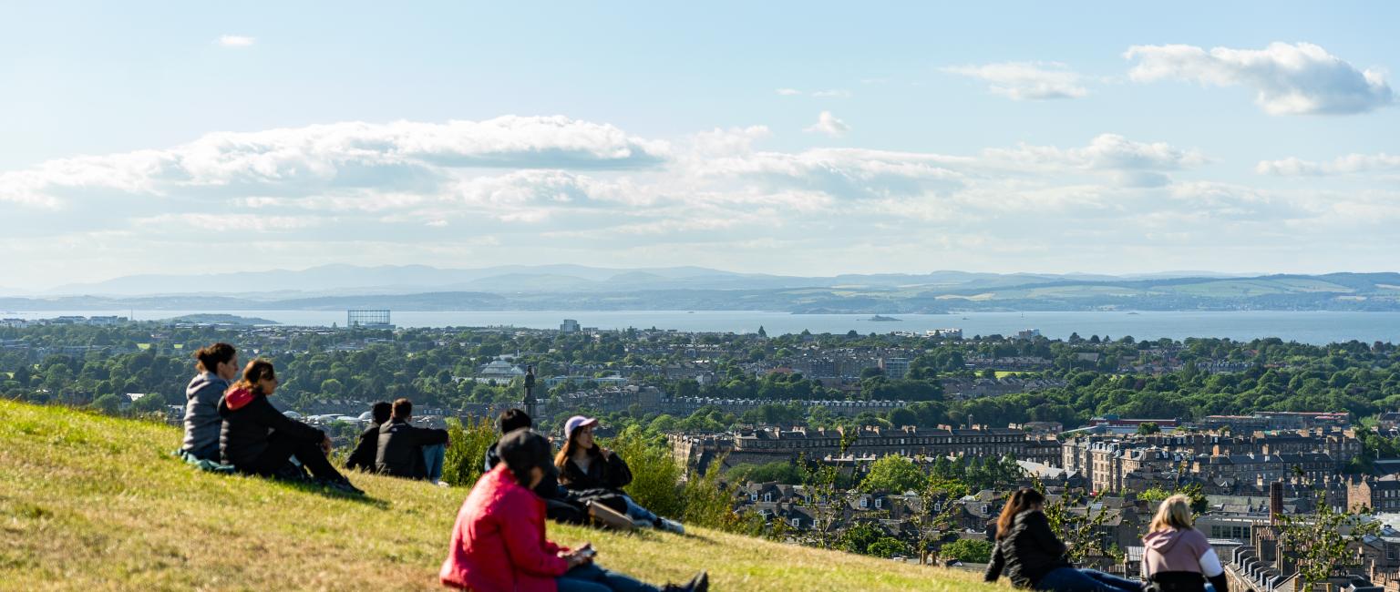 People stretched out on the grass of a mountain hill watching the views of the city of Edinburgh, UK.