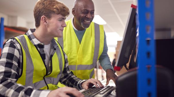 Intern With Supervisor Working In Busy Modern Warehouse On Computer