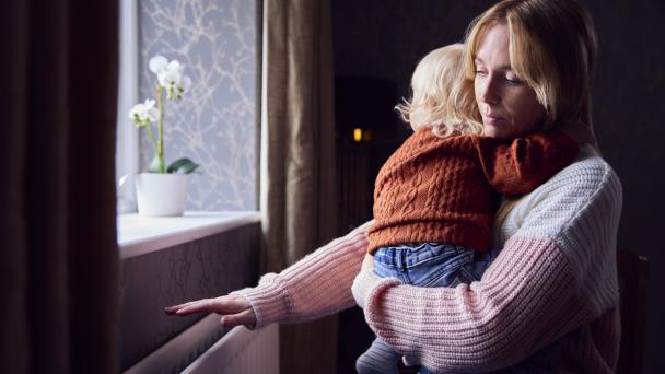 Mother With Son Trying To Keep Warm By Radiator At Home During Cost Of Living Energy Crisis 