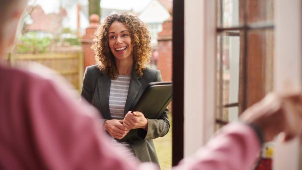 Unidentifiable person opening their front door in the foreground, and a woman with folder working as a survey interviewer smiling. 