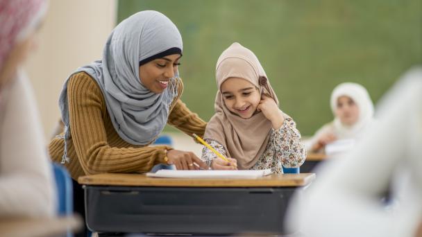 A muslim girl smiles as she sits in class learning. She is looking at her paper that her teacher is pointing at.