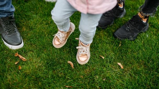 Image showing a family from above so only their legs and feet are visible. Parents and one child, standing on grass.