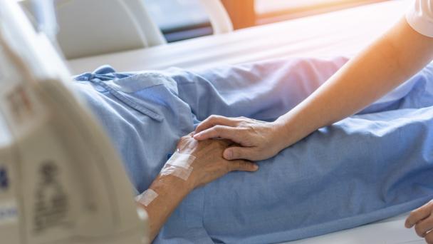 Caregiver holding elderly senior patient (ageing old adult person) hand in hospital bed.