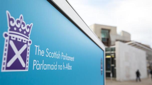 A blue sign to the Scottish Parliament, Parlamaid na h-Alba, is in the foreground and background is blurred buildings with a person walking in front.