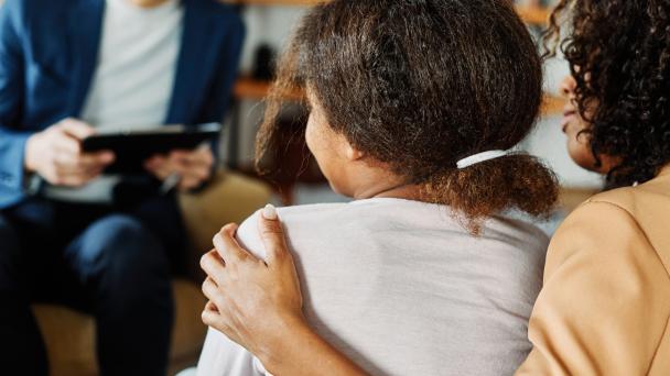 Mother has her arm around her child in the foreground, talking to a therapist, advisor or support worker in the background.