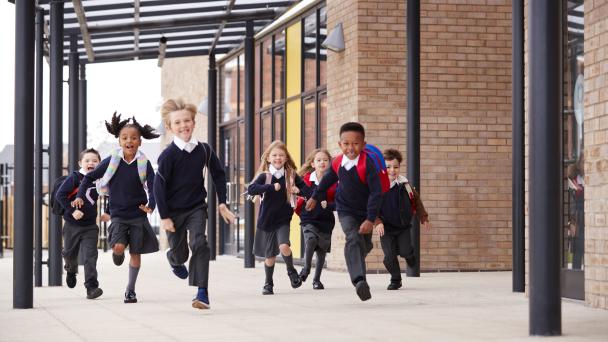 Primary school kids, wearing school uniforms and backpacks, running on a walkway outside their school building, front view