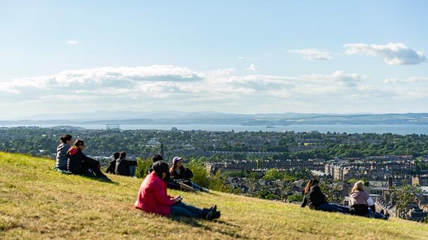 People stretched out on the grass of a mountain hill watching the views of the city of Edinburgh, UK.