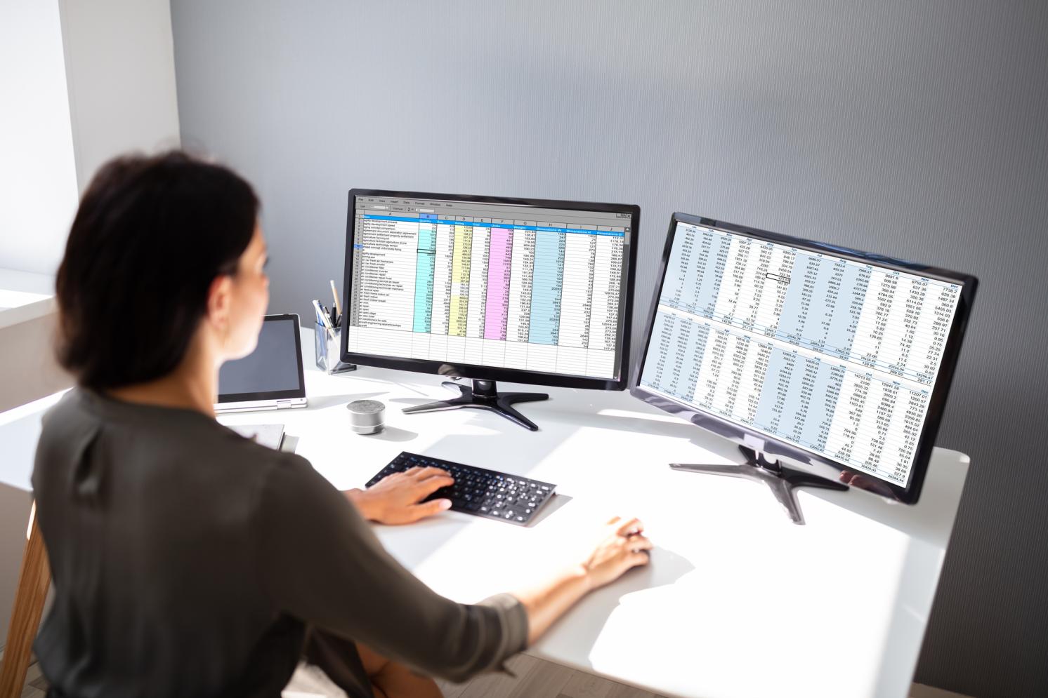 Female looking at two computer screens conducting analysis of spreadsheets