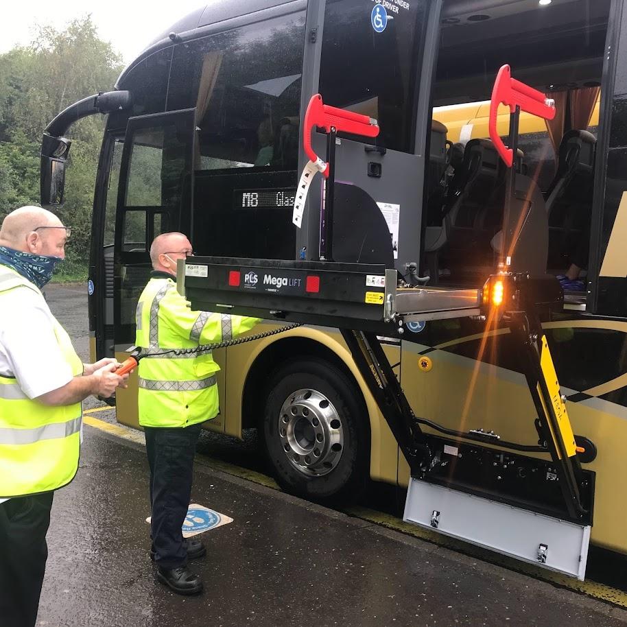 Two men operate a lift attached to the side of a coach. The lift looks like a large platform and it is currently parallel to the coach floor.