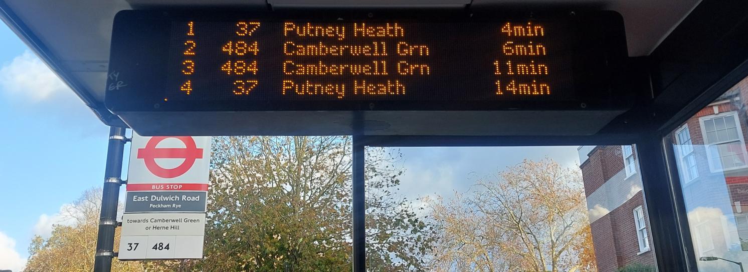 A digital display hangs from the top of a bus shelter. The display lists the bus number, destination and expected arrival time for the next four buses that will arrive at the bus stop.